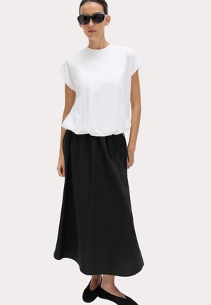 Femme portant de grandes lunettes de soleil noires, un haut blanc à manches courtes, une longue jupe noire et des ballerines noires, debout devant un fond clair uni.