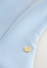 Gaiši zils audums ar mazu apaļotu zelta piekariņu, uz kura ir gravēta burts "B", piešūts ar gaiši zilu diegu.