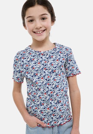 Fille souriante aux cheveux foncés portant un haut à manches courtes à motif floral avec des bords rouges, la main dans la poche d’un pantalon rayé, sur fond uni.