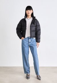 Svart pufferjakke med høy krage og brodert logo, brukt over en lys grå T-skjorte. Blå løst sittende jeans og svarte hælede sko.