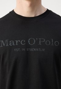 Melna kokvilnas T-kreklu ar apaļu kakla izgriezumu un īsām piedurknēm. Priekšpusē ir prints: "Marc O’Polo EST. IN STOCKHOLM" smalkā pelēkā krāsā.