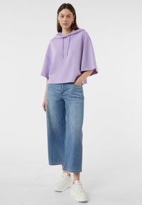 Fioletowy oversized sweatshirt z regulowanym kapturkiem, połączony z jasnoniebieskimi spodniami o szerokich nogawkach i białymi sneakersami, na jednolitym tle.