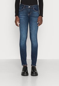 Jeans de denim azul de corte Slim fit en una persona de pie que lleva mocasines negros y una blusa negra de manga larga, contra un fondo blanco.
