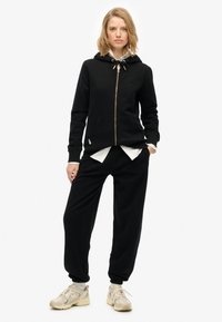 Felpa nera con zip e cappuccio regolabile, tasche frontali e vestibilità comoda, abbinata a pantaloni della tuta neri e sneaker beige.