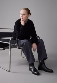 Bottines en cuir noir avec un bout carré et une fermeture éclair latérale, associées à un pantalon en denim gris ample et un pull noir, assis sur une chaise moderne.