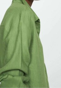 Camisa de lino verde con un corte relajado, que presenta un cuello y mangas enrolladas, exhibida sobre un fondo liso.