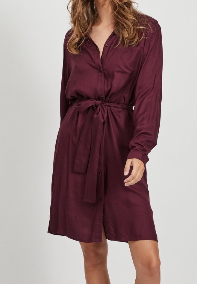 Femme portant une robe chemise bordeaux à manches longues, longueur au genou, boutonnée et avec une ceinture nouée à la taille, sur un fond uni.