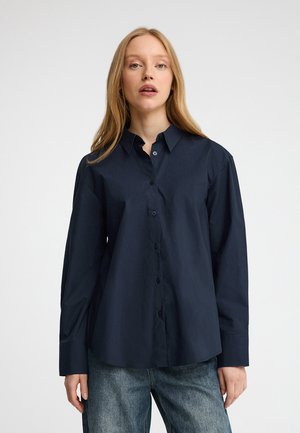 Mujer con cabello largo y rubio que lleva una camisa azul marino oscuro abotonada y vaqueros azules, de pie frente a un fondo blanco.