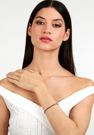 Bracciale in argento con gemme colorate, con un design delicato sul polso sopra un vestito bianco off-shoulder con dettagli in pizzo.