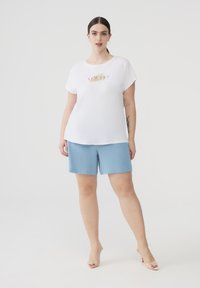 T-shirt bianca in cotone con grafica "ALWAYS", abbinata a pantaloni corti azzurri testurizzati. Il modello indossa sandali con tacco trasparente. Design semplice e casual.