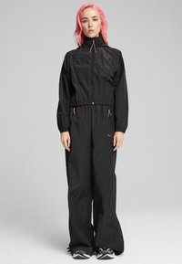 Veste coupe-vent noire avec col haut, poignets élastiques, taille cintrée et poches zippées. Tissu léger et texturé avec des accents orange contrastants.