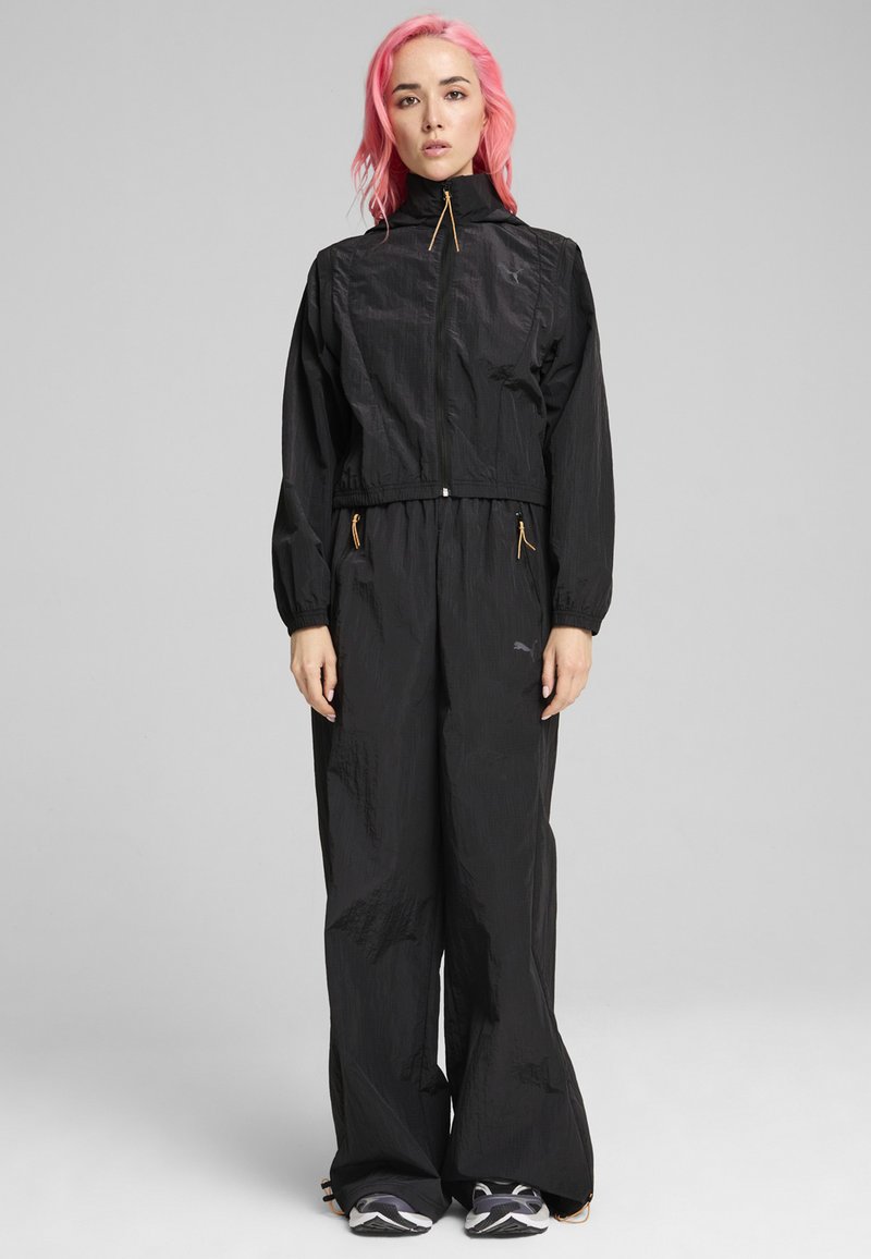 Veste coupe-vent noire avec col haut, poignets élastiques, taille cintrée et poches zippées. Tissu léger et texturé avec des accents orange contrastants.