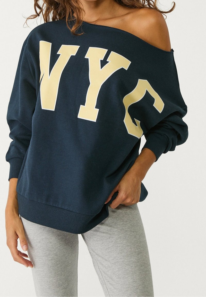 Femme portant un sweat-shirt bleu marine épaules dénudées avec de grandes lettres « NYC » jaune pâle et des leggings gris clair, main posée sur la hanche.