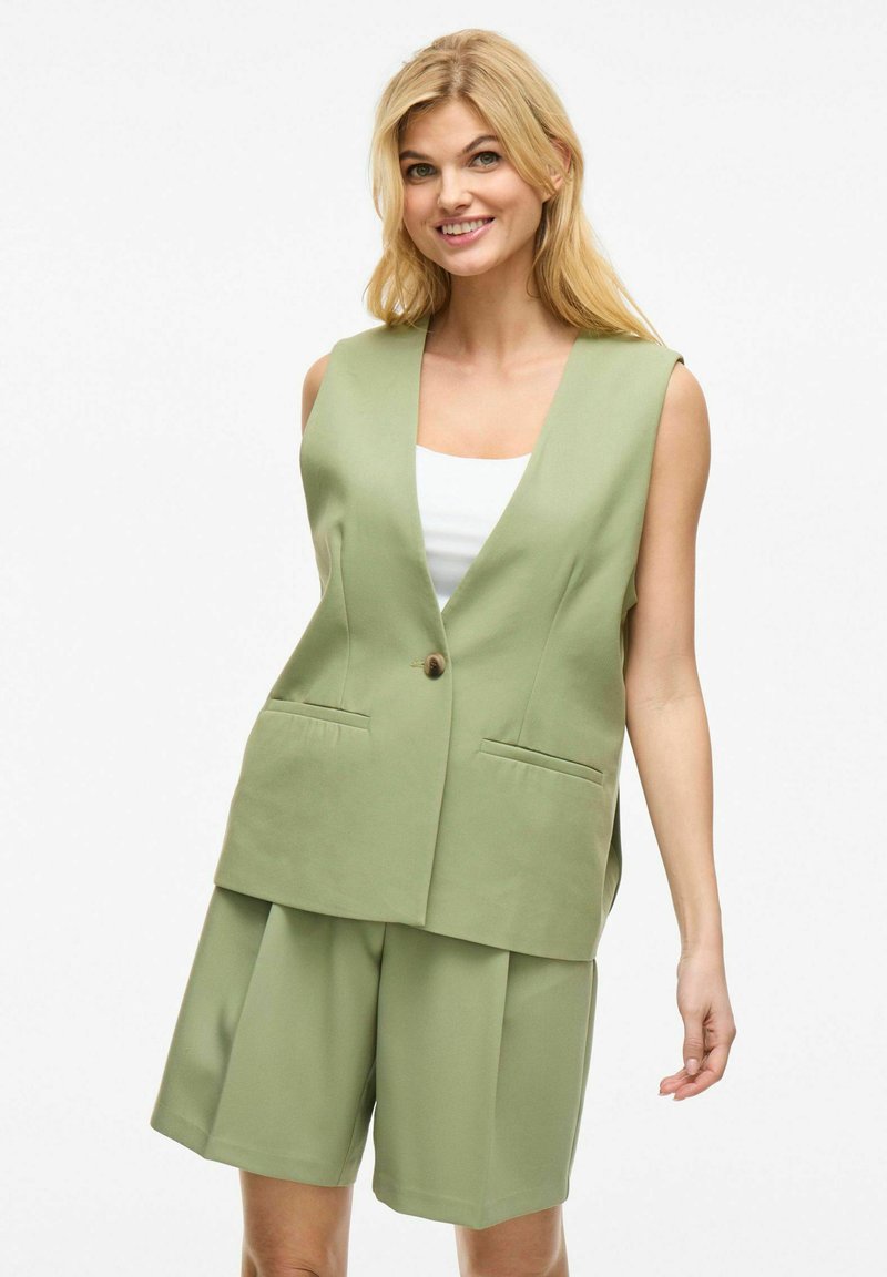 Blazer sin mangas verde con un solo botón, dos bolsillos frontales y un corte relajado, combinado con pantalones cortos holgados a juego.