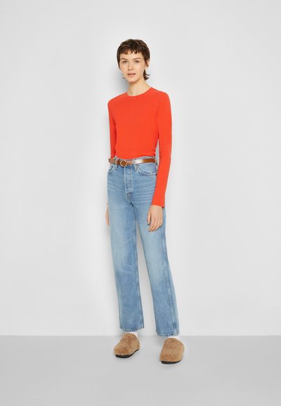 Camisa vermelha de mangas longas, jeans de cintura alta de tom azul claro, cinto prateado com fivela circular e pantufas fofy bejes. Design minimalista, ajuste casual.