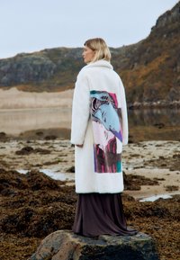 Vit pälsrock med färggrafik på ryggen, över en lång brun klänning, framför en naturlandskap.