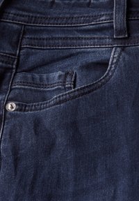 Ciemne dżinsy o gładkiej teksturze, z miedzianymi nitami oraz małą kieszonką na monetę z przodu. Czyste szwy podkreślają design.