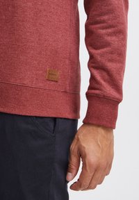 Sweatshirt rouge en tissu texturé, avec des poignets côtelés et un patch en cuir marron sur le côté inférieur, associé à un pantalon sombre.