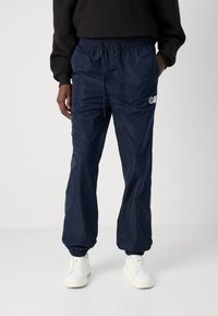 Navyblauwe, lichtgewicht, waterdichte broek met elastische tailleband en boorden; heeft een logo in het wit op het linkerbeen. Draag ze met witte sneakers.