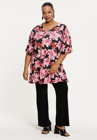 Bloemige tuniek met korte mouwen, met roze en rode bloemen op een zwarte achtergrond. Gecombineerd met zwarte wijde broek en zwarte sandalen met blokhakken.