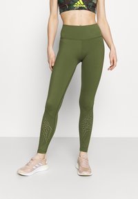 Oliwkowe legginsy z wysokim stanem, z teksturowanym wzorem na dolnej części nóg i małymi wycięciami, w połączeniu z jasnoróżowymi butami sportowymi.