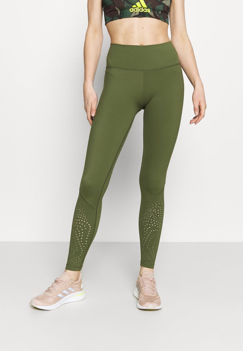 Oliwkowe legginsy z wysokim stanem, z teksturowanym wzorem na dolnej części nóg i małymi wycięciami, w połączeniu z jasnoróżowymi butami sportowymi.
