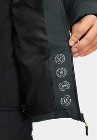 Wnętrze czarnej kurtki izolacyjnej, pokazujące teksturę materiału i cechy wodoodporne. Zawiera drukowane ikony dotyczące oddychalności i materiałów z recyklingu.