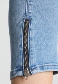 Primer plano de una pierna de pantalón de mezclilla azul claro con una cremallera metálica vertical parcialmente desabrochada cerca del dobladillo, revelando la piel debajo.