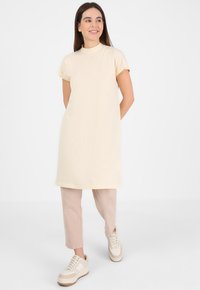Abito in cotone beige con collo alto e maniche corte, abbinato a pantaloni rosa chiaro e sneaker bianche con dettagli color cammello.