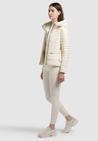 Femme debout portant une veste matelassée crème à capuche, des leggings assortis et des baskets de couleur claire sur un fond gris clair uni.