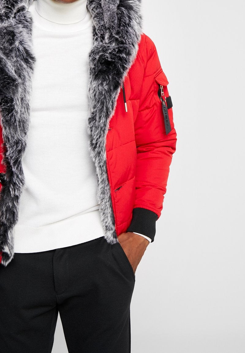 Rote Steppjacke mit grauem Kunstfellkragen, Reißverschlusstasche am Ärmel und gerippten Bündchen, kombiniert mit einem weißen Rollkragenpullover und schwarzen Hosen.