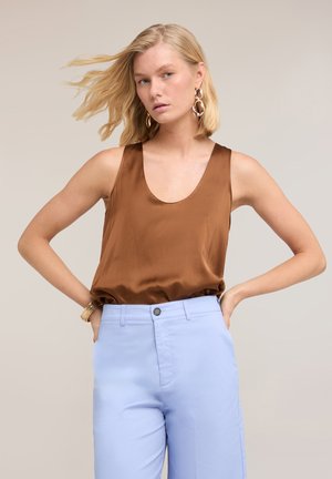 Vrouw met blond haar, grote gouden oorbellen, een bruin mouwloos topje en lichtblauwe broek met hoge taille, poserend met handen in de zij.