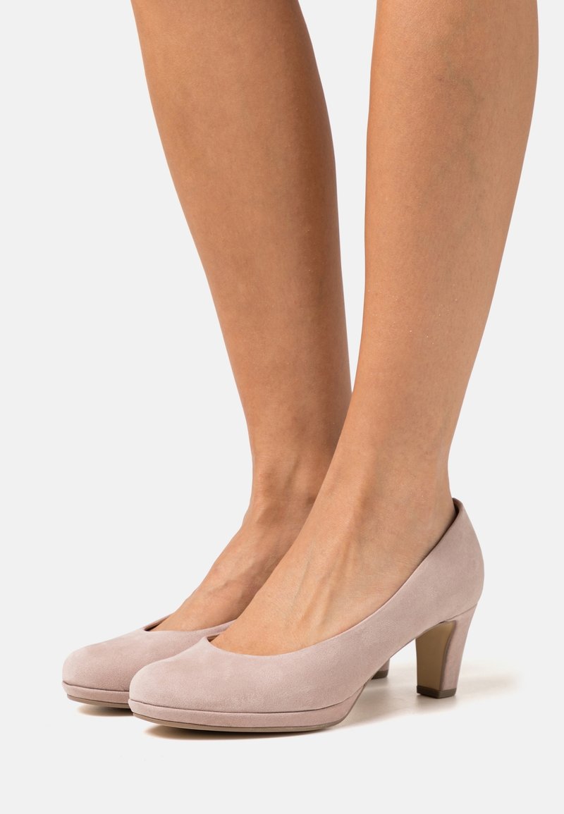 Paire d'escarpins en daim rose clair à talons moyens portés pieds nus sur un fond blanc uni.