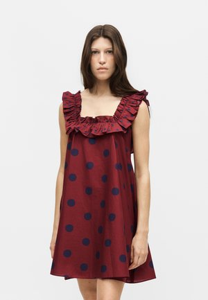 Fiatal nő hosszú barna hajjal, ujjatlan bordó ruhát visel tengerészkék pöttyökkel és fodros, négyzet alakú nyakkivágással, egy egyszínű háttér előtt áll.