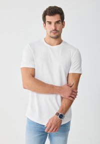 Camiseta blanca de algodón con cuello redondo y mangas cortas, combinada con jeans de mezclilla azul claro. Presenta un reloj marrón en la muñeca.