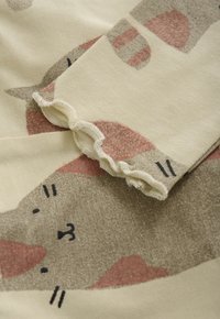 Crèmekleurige stof met een speels kattenpatroon in beige en roze, met gerimpelde randen op een opgevouwen mouw of manchet.