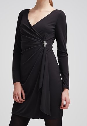Woman wearing a black long-sleeve V-neck dress with draped detail and a decorative brooch at the waist.