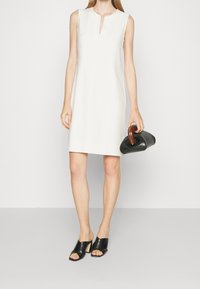 Robe blanche sans manches avec un col en forme de goutte, texture lisse, coupe droite, associée à un sac à main noir et des mules noires ouvertes à l\'avant.