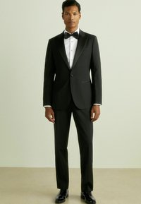 Costum negru din tuxedo cu rever din satin, cămașă albă, papion negru și pantofi negri lucioși. Croială adaptată și linii curate.