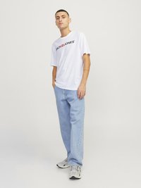 Balta trumpa rankovėmis marškinėliai su juodu "JACK & JONES" tekstu, derinami su šviesiai mėlynomis tiesiomis džinsomis ir pilkais sportbačiais.