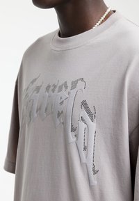 Homme portant un t-shirt oversize gris clair avec un motif de texte gothique à pois noirs et un collier de perles sur fond blanc.