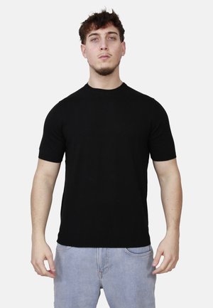 Giovane uomo con capelli corti e ricci, indossa una maglietta nera semplice a maniche corte e jeans azzurri chiari, in piedi davanti a uno sfondo bianco.