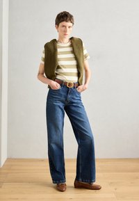 Striped olive and cream knit tank top layered over a white shirt, paired with high-waisted blue jeans and brown loafers. Wooden floor background.
