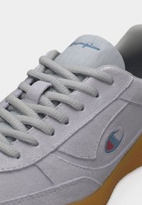 Κοντινό πλάνο γκρι αθλητικού παπουτσιού από σουέντ με γκρι κορδόνια, σόλα από καουτσούκ και το λογότυπο της Champion στο πλάι και στη γλώσσα.