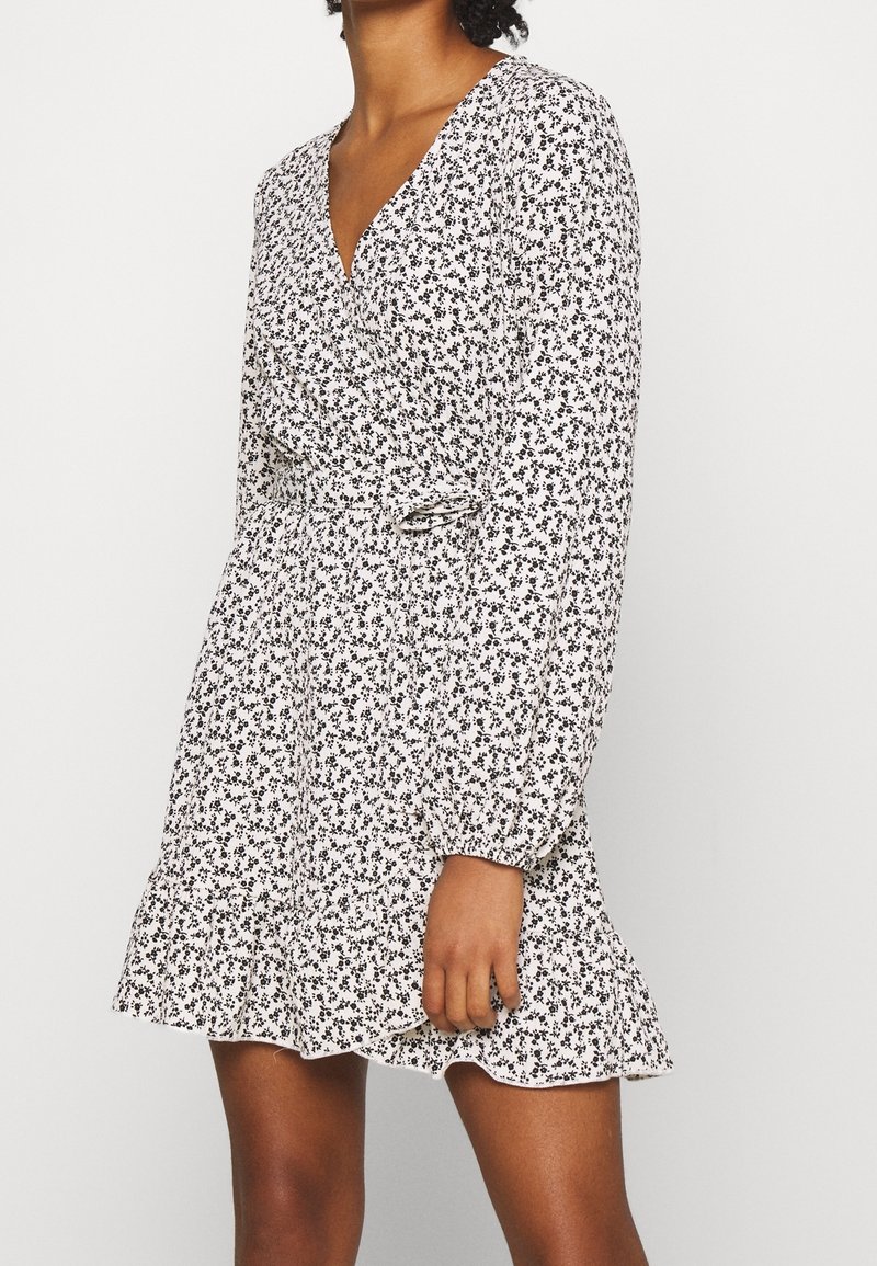 Femme portant une robe blanche à manches longues avec imprimé floral noir et ourlet volanté, debout devant un fond clair uni.