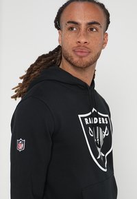 Felpa nera con cappuccio realizzata in tessuto morbido, caratterizzata da un grande logo dei Raiders in bianco. Include un patch della NFL sulla manica sinistra e una tasca a marsupio.