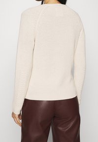 Femme portant un pull en maille texturée crème et un pantalon en cuir marron foncé, vue de dos, sur un fond clair uni.