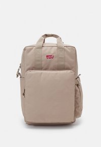 Μπεζ σακίδιο με δύο επάνω λαβές, κύριο διαμέρισμα με φερμουάρ, μπροστινή τσέπη και πλαϊνή τσέπη από δίχτυ. Διαθέτει κόκκινο λογότυπο Levi's μπροστά.