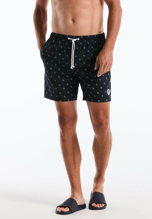 Man draagt zwarte zwemshorts met witte ankerpatronen en een wit trekkoord, gecombineerd met zwarte slippers met reliëflogo's.