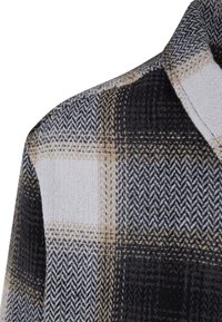 Tkanina w kratę w kolorach czarnym, białym i beżowym o teksturowanym wykończeniu; zawiera wzór w jodełkę oraz detal kołnierza. Zbliżenie na krawędź odzieży.